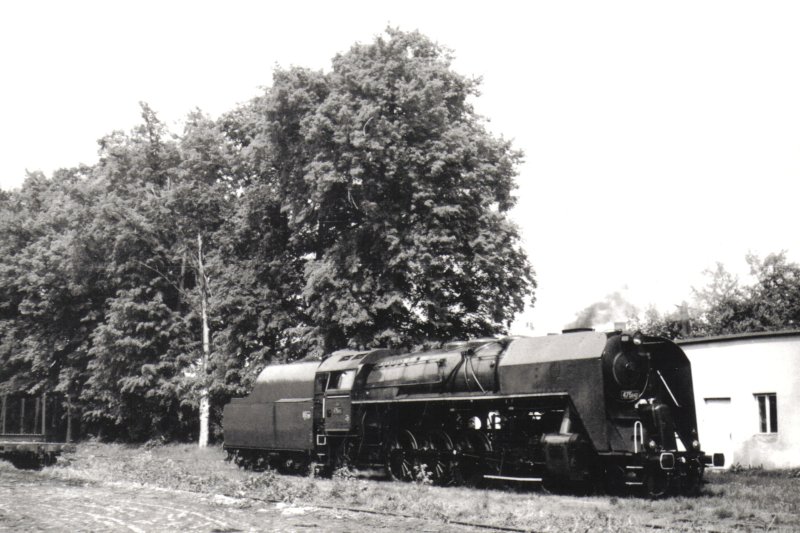  Parný rušeň 475.1142 v žst. Zásmuky. Pohľad zprava zpredu. Vzadu sklad a stromy. Anon., 1.6.1986. 180 x 130 