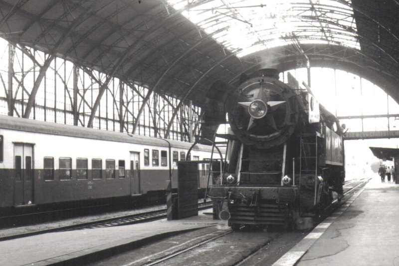  Parný rušeň 477.043 v hale žst. Praha hl n. Pohľad zpredu. Vľavo jednotka EM 475.1. Anon., cca 1985. 125 x 90 