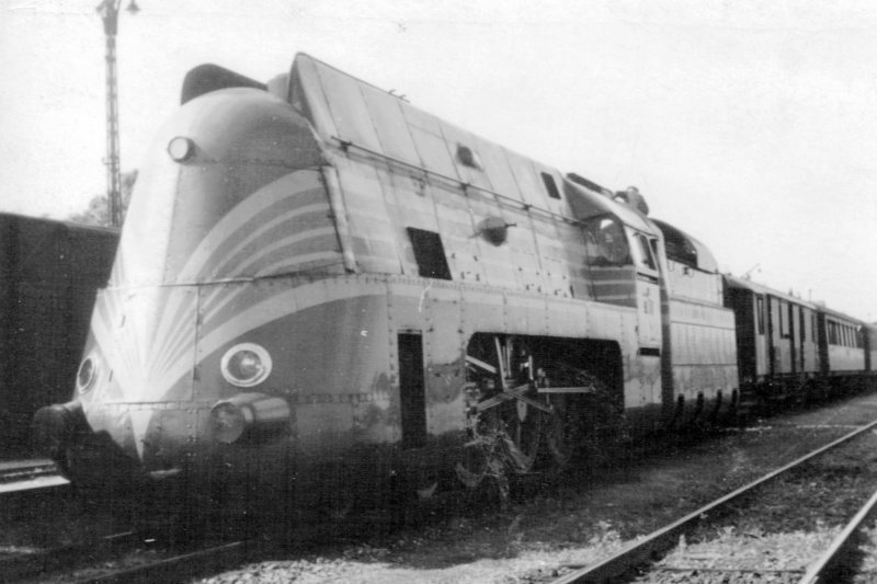  Parný rušeň 386.001 SŽ + 1 Da + súprava podvozkových vozňov v koľajišti stanici. Vzadu osvetľ. stožiar. Anon., cca 1940. 108 x 78 