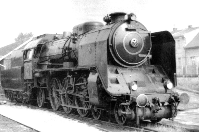  Parný rušeň 387.043 v stanici Nymburk-město. Pohľad zprava zpredu. Vzadu obyt. budovy. Anon., 17.9.1982. 122 x 84 