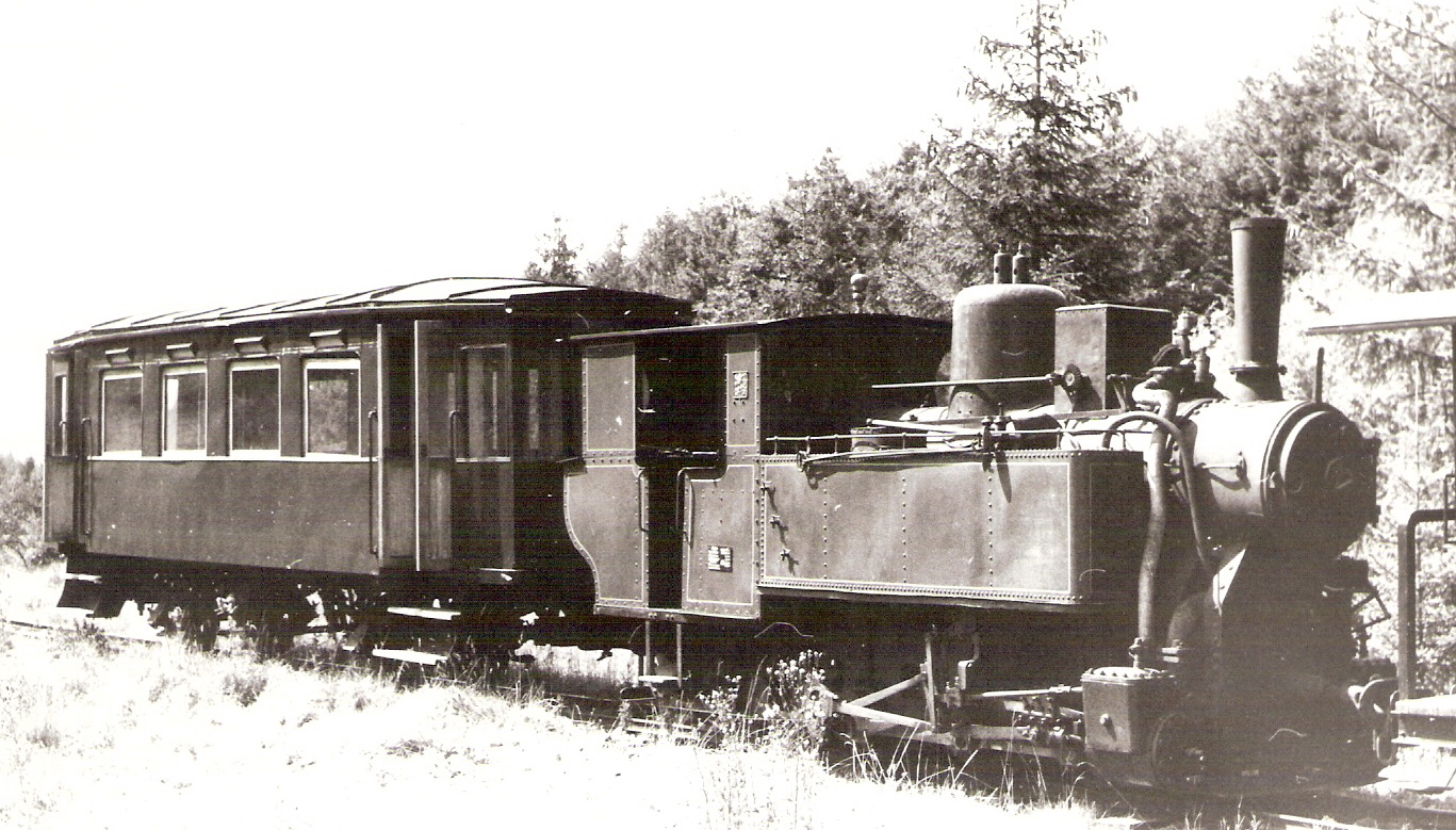  Úzkorozch. parný rušeň štvornápravový MÁV + tramv. príves. dvojnápr. vozeň na ZLŽ. Pohľad zprava zpredu. Vzadu zalesnený svah. Anon., cca 1980. 178 x 110 