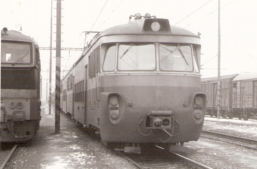  Elektrická motorová jednotka EM 475.2015 v žst. Týniště nad Orlicí. Pohľad zpredu. Vľavo T 478.3?85, vpravo nákladné vozne. Trolej. Foto: J. Adolf, 1.3.1985. 141 x 100 