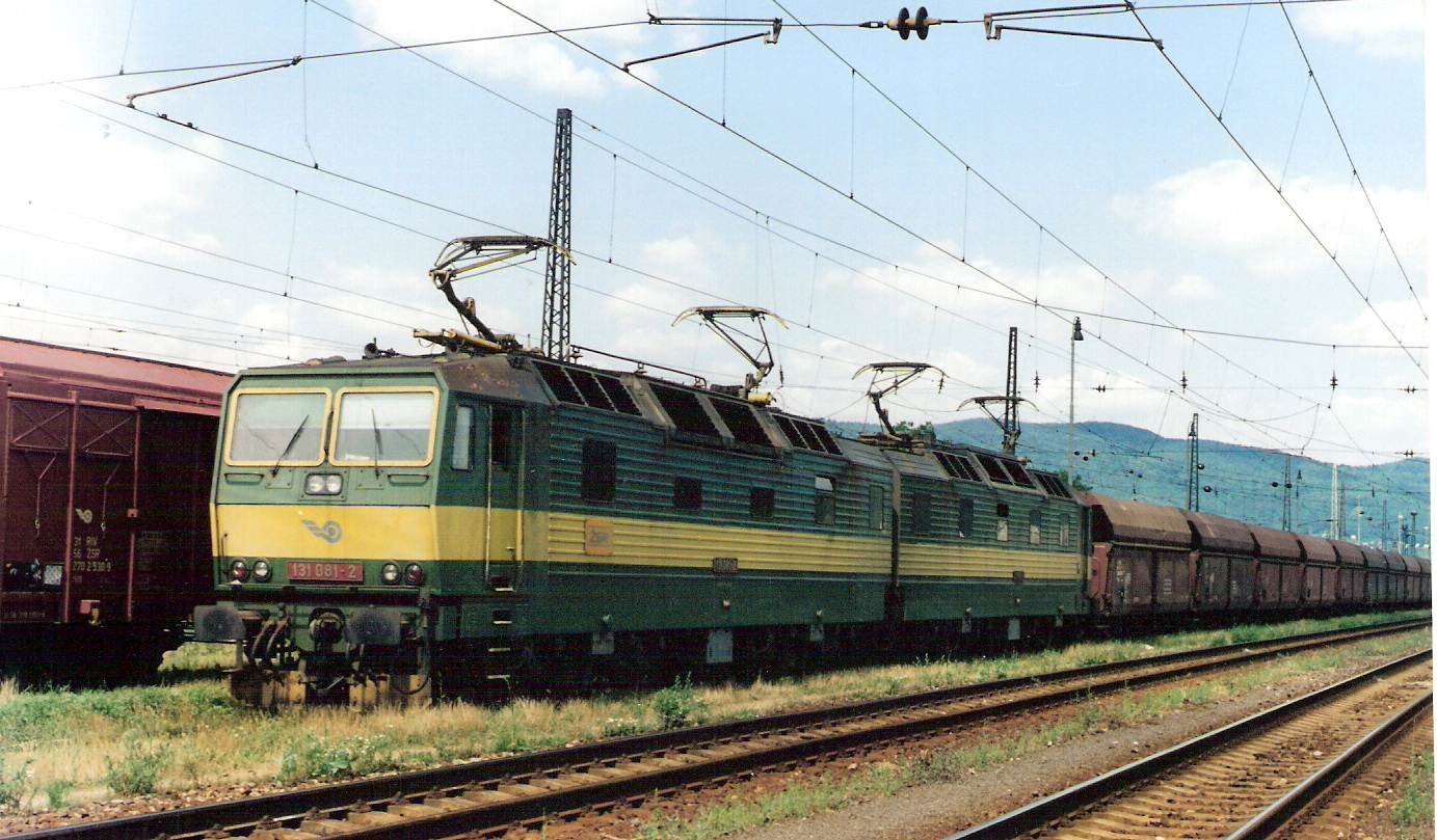  Elektrický rušeň 131 081-2 so súpravou Wap v žst. Púchov. Pohľad predobočný. Vľavo vozeň Za/Has. Trolej, osvetľ. stožiar. vzadu hory. Foto: M. Entner, jún 1997. 178 x 126, COLOR 