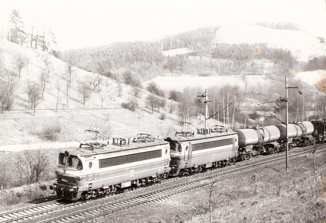  Dva elektrické rušne S 489.0 (prvý 080) ťahajú nákladný vlak na dvojkoľajnej trati v horskom údolí. Vzadu kopec s lúkami a lesom. Foto: Anon., cca 1985. 178 x 130 