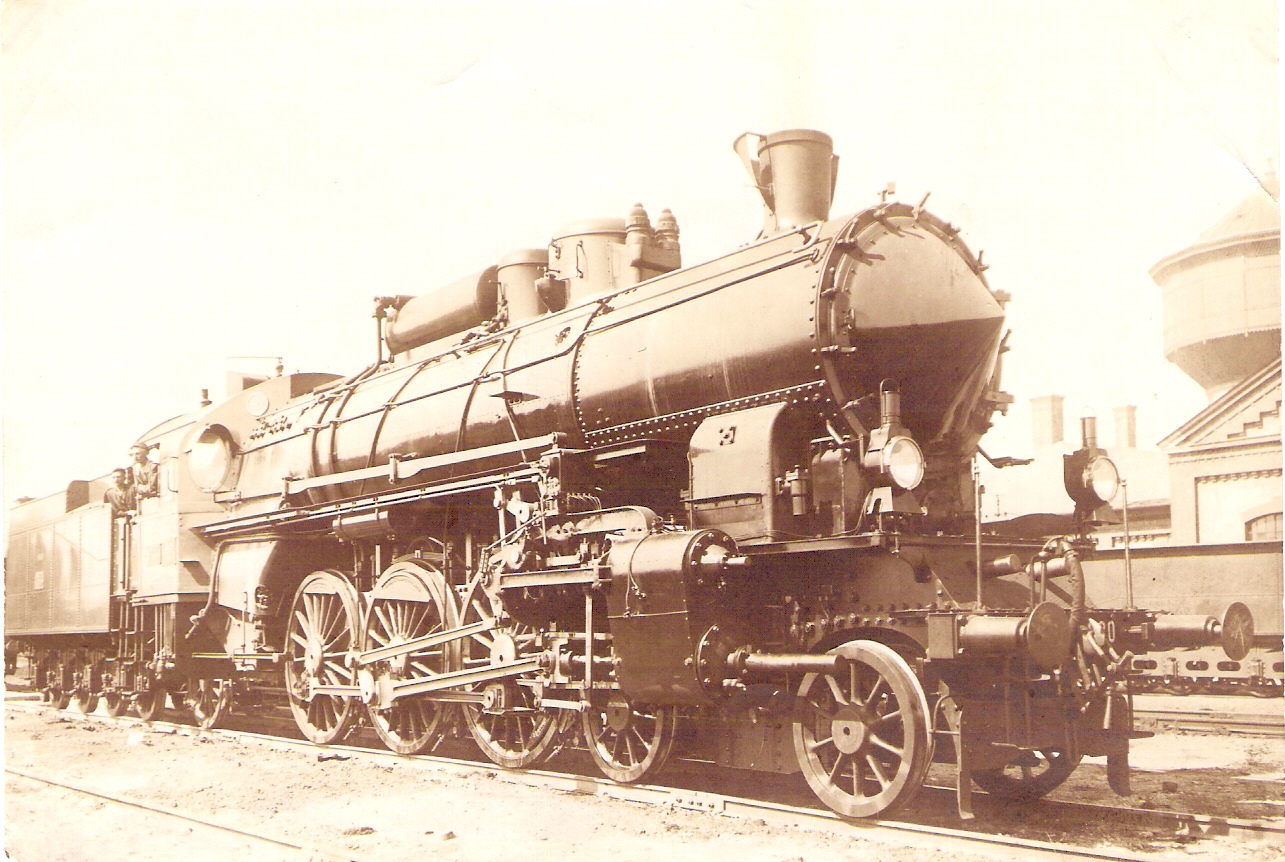  Parný rušeň 301 MÁV v depe. Pohľad zprava zpredu. Vpravo vzadu tender iného parného rušňa, vodáreň a dielenské budovy. Foto: Anon., cca 1914. 162 x 118 