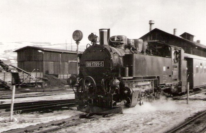  Úzkorozch. parný rušeň 99 1791-5 DR pri odchode zo žst. Oberwiesenthal. Pohľad zľava zpredu. Vpravo drevená remíza, vľavo hodiny, vodný žeriav a drevená hala. Foto: Anon., 12.2.1982. 123 x 83 