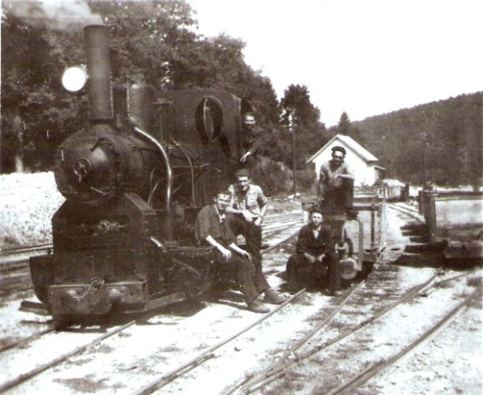  Úzkorozch. par. rušeň (zrejme 3-nápr. MÁV s rovným komínom) na zhl. veľk. koľajiska. Pohľ.zľava zpredu. Pri rušni pózujú piati muži v pracovnom. Vzadu malé repné drev.huntíky a murov. budova. Foto: Anon., cca 1960. 90 x 90 