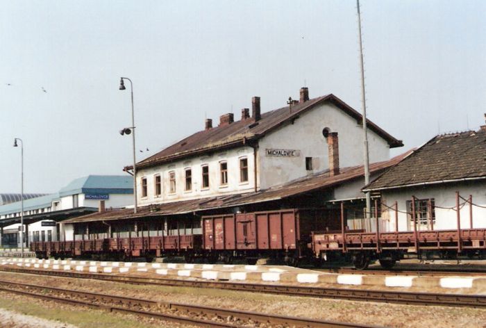  Michalovce - výpravná budova. Šikmý pohľad zo strany koľají od Košíc. Osvetľovacie stožiare, pred budovou nákladné vozne. Vľavo nová výpravná budova. Foto: K. Kočiško, cca 2000. 125 x 88, COLOR 
