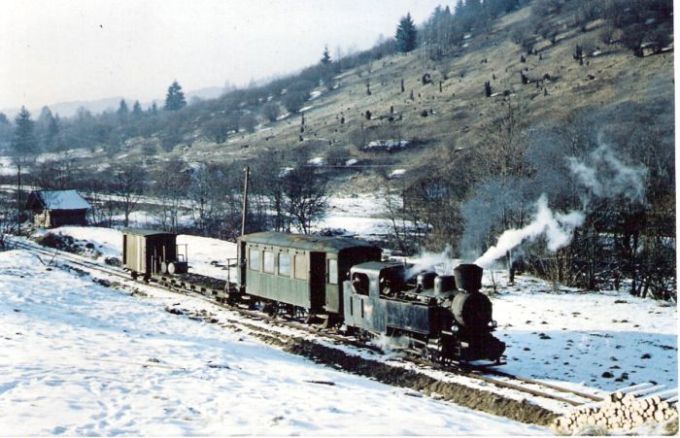  Vlak Vígľaš.lesnej žel. s par.rušňom C 760/90d, tramv. prívesom, plošovým a malým prac. vozňom. Zimný pohľ.zprava zpredu. Vpravo stráň s riedko roztrús. stromami. Telegrafné tyče, vľavo drevenica. Foto: Anon., cca 1970. 153 x 100 