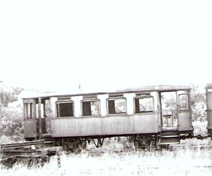  Tramvajový dvojnápravový príves Vígľašskej lesnej železnice. Pohľad zboku. Vľavo koniec oplenového podvozka. Vzadu obytné domy. Foto: V. Hlávek, cca 1975. 63 x 43 