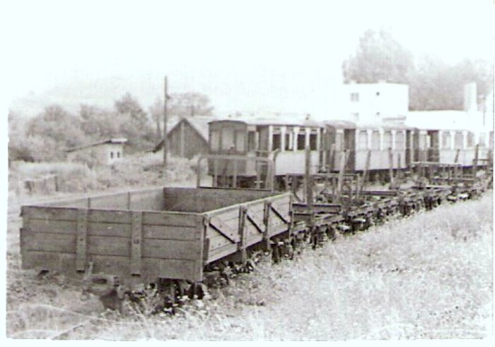  Vígľašská lesná železnica - stanica Vígľaš. V popredí dvojnápr. nízkostenný vozeň, za ním plošinové podvozky, vzadu tri tramvaj. prívesy. Za nimi vpravo obytné domy a hora. Foto: V. Hlávek, cca 1975. 60 x 43 