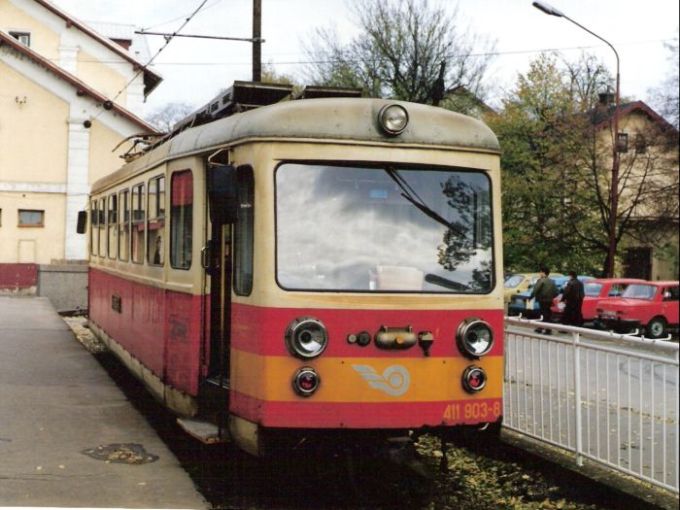  Elektrický motorový vozeň 411 903-8 v Trenčianskej Teplej. Pohľad predobočný. Vľavo bočné priečelie výpravnej budovy, vpravo os. autá a železn. Kolónia. Foto: M. Entner, 25.10.1995. 178 x 126 