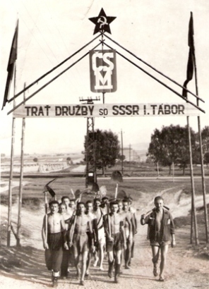  Skupina ľudí v pracovnom v tvare s nástrojmi pod bránou s nápisom Trať Družby so SSSR I. tábor, znakom ČSM a hviezdou s kosákom a kladivom. Anon., cca 1950. 175x125 