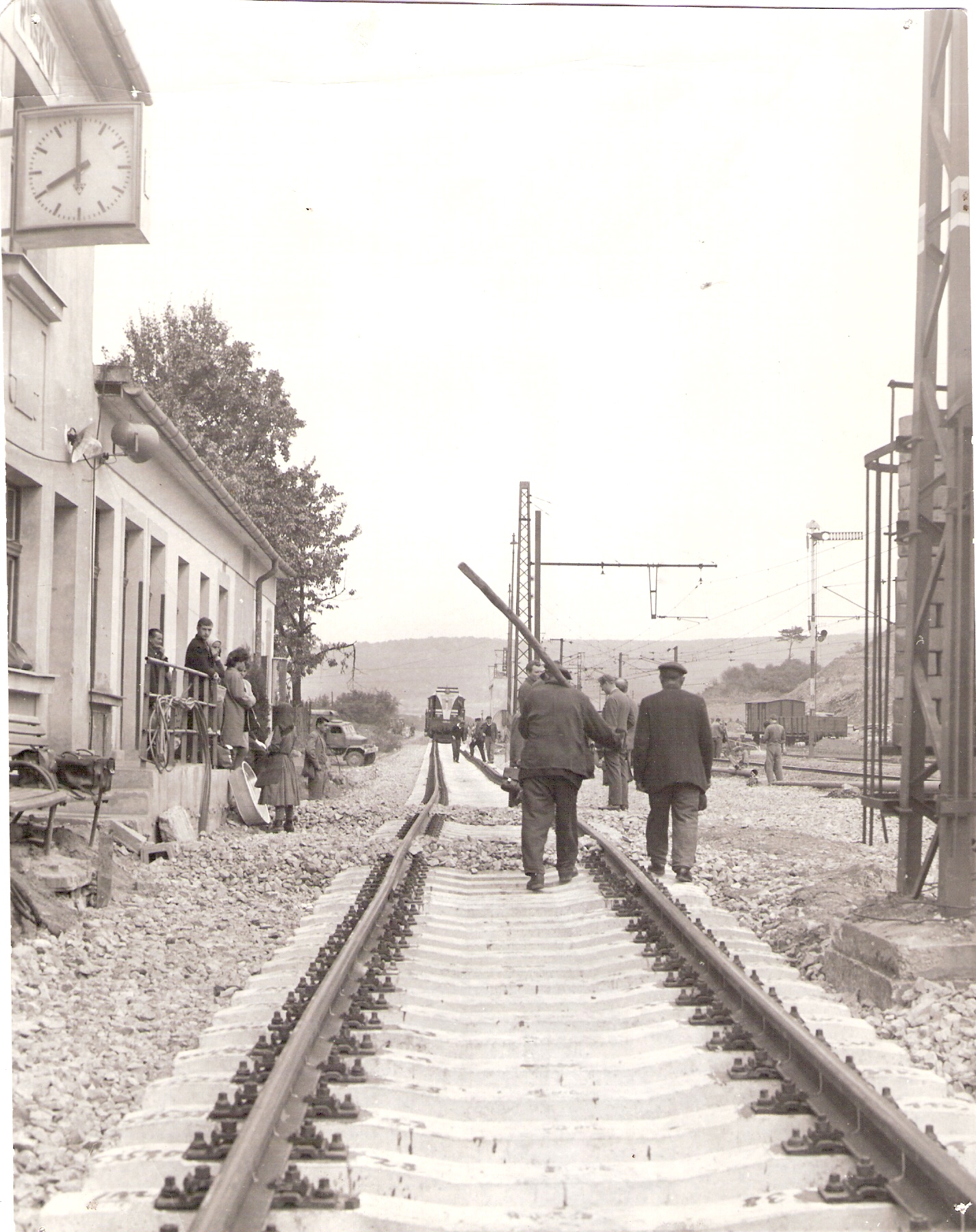  Ruskov - koľajisko počas výstavby ŠRT. Pohľad v osi novej nepodbitej koľaje, na nej vzadu T 458.5. Vľavo priečelie výpravnej budovy, vedľa P V3S. Vpravo staré koľajisko s mech. náv., s trolejmi. Robotníci, civilisti. Foto: Anon, cca 1965. 180 x 229 