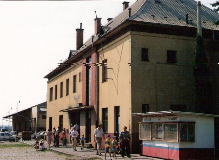  Bánovce nad Bebravou - výpravná budova. Šikmý pohľad z predstaničného priestoru od Trenčína. Vpredu stánok PNS, vľavo sklad. Ľudia. Foto: M. Entner, 25.8.1996. 125 x 90, COLOR 