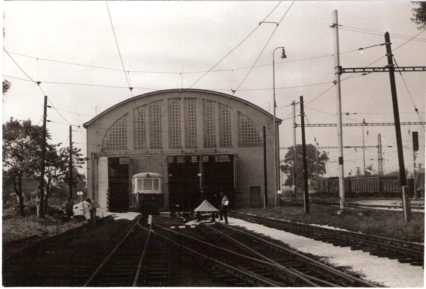  Trenčianska Teplá - elektrické depo nové. Čelný pohľad v osi koľaje od Žiliny. Pred depom prívesný vozeň. Ľudia pri práci. Foto: Anon., cca 1985. 178 x 120 