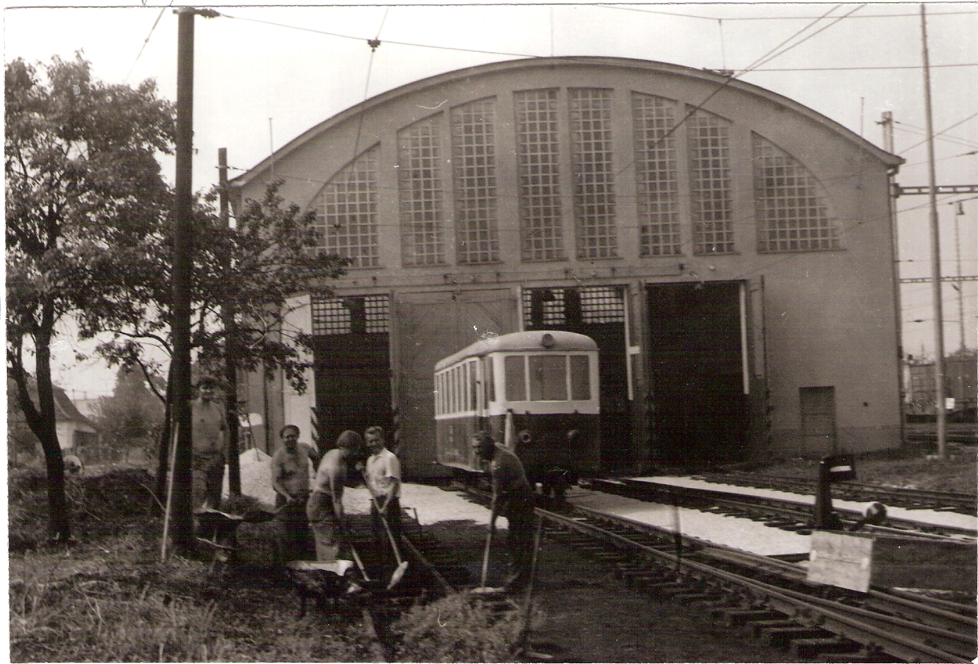  Trenčianska Teplá - elektrické depo nové. Čelný pohľad v osi koľaje od Žiliny. Pred depom prívesný vozeň. Ľudia pri práci. Foto: Anon., cca 1985. 178 x 120 