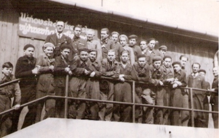  Dospelý muž v civile so skupinou učňov v pracovnom pred drevenou budovou s nápisom Učňovské stredisko výhrevne Bratislava. Anonym, cca 1950. 120 x 85 
