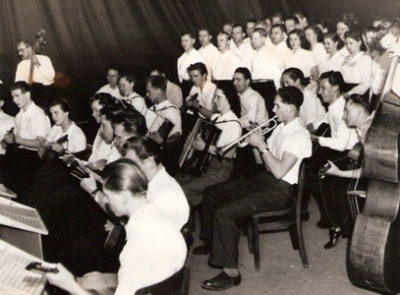  Estrádny orchester a zmiešaný spevácky zbor počas vystúpenia. Text: Trať Družby - Spišské Vlachy. Anon., cca 1950. 164 x 122 