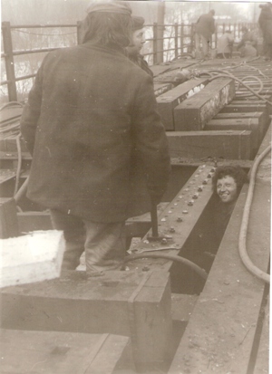  Skupina robotníkov (jednému trčí iba hlava) pri oprave oceľovej konštrukcia mosta nitovaním (zrejme Červený most v Bratislave). Foto: Macura, M., cca 1980. 180 x 130 