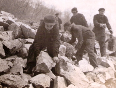 Skupina mužov a jedna žena v pracovnom s nápisom ČSD a znakom SSM prenášajú kamene. Text: Stavba Trate 1. mája Modrý Kameň - Szakal. Anon., cca 1955. 160 x 120. 