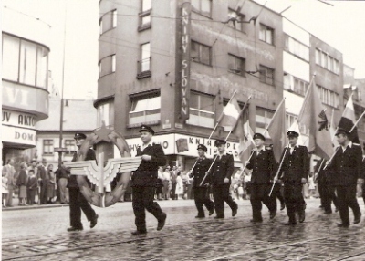  Skupina mužov v železničných rovnošatách s vlajkami a znakom okrídleného kolesa v prvomájovom sprievode, Bratislava, pred predajňou obuvi pri Michalskej bráne. Anonym, cca 1960. 115 x 85 