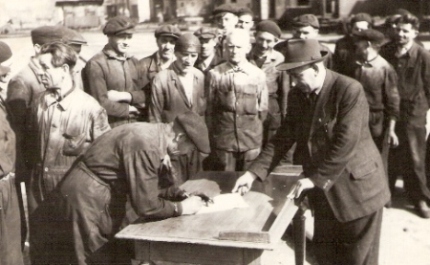  Skupina mužov v pracovnom, jeden v civile, pri podpise dokumentu na stole. V pozadí areál dielní s os. vozňami. Text: Dielne ČSD Vrútky, súdruhovia z vozovky podpisujú soc. záväzky, prítomný je prednosta dielní J. Gíreth. Kľučka, cca 1950. 127 x 80 