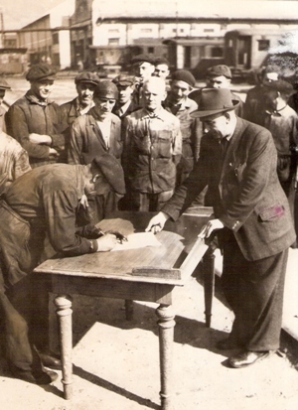  Skupina mužov v pracovnom, jeden v civile, pri podpise dokumentu na stole. V pozadí areál dielní s os. vozňami. Text: Dielne ČSD Vrútky, súdruhovia z vozovky podpisujú soc. záväzky, prítomný je prednosta dielní J. Gíreth. Kľučka, cca 1950. 170 x 127 