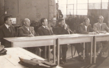  Skupina mužov v civile sediacich za stolom, za nimi zadná časť autobusu. Text: Dielne ČSAD. Anonym, cca 1950. 155 x 105 