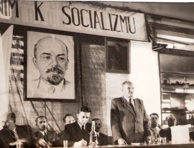  Skupina mužov v civile sediacich za stolom, jeden prednáša prejav. V pozadí obraz Lenina a Stalina. Text: Dielne ČSAD. Anonym, cca 1950. 185 x 138 