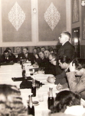  Skupina ľudí v civile sediacich za stolom, jeden prednáša prejav. Text: Ústredný riaditeľ s. Bezek pri prejave na pracovnej porade na Vrútkach. Anonym, cca 1950. 113 x 83 