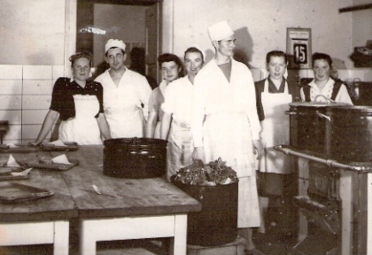  Skupina ľudí v bielych plášťoch a v civile v prostredí závodnej kuchyne. Text: Železničná kuchyňa. Anonym, cca 1950. 120 x 85 