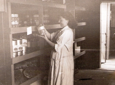  Žena v bielom plášti vyberá z regálu v skladovom vozni konzervu. Text: Železničná kuchyňa. Anonym, cca 1950. 138 x 112 