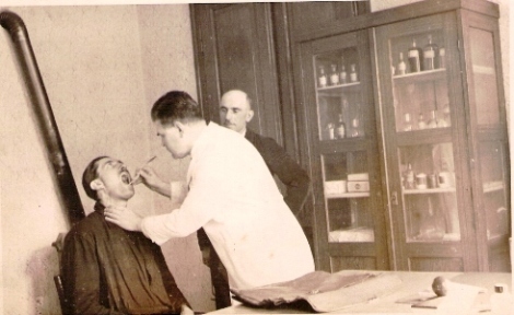  Lekár pri zubnej prehliadke muža v pracovnom odeve. Prizerá sa muž v civile. V pozadí kachle, dvere a drevená lieková skriňa. Anonym, cca 1950. 130 x 80 