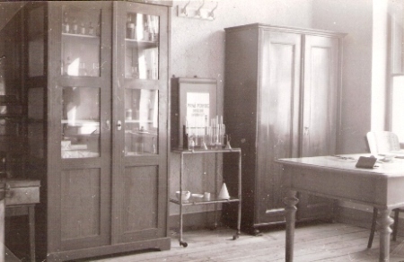  Interiér zdravotnej ambulancie s drevenou liekovou skriňou, inou drevenou skriňou, vešiakom a dreveným stolom. Anonym, cca 1950. 138 x 90 