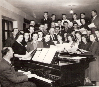  Zmiešaný spevácky zbor s klavíristom (všetci v civile). Text: Spevokol železničiarov na Oblastnom riaditeľstve v Bratislave. Foto: Kľučka, cca 1950. 122 x 125 