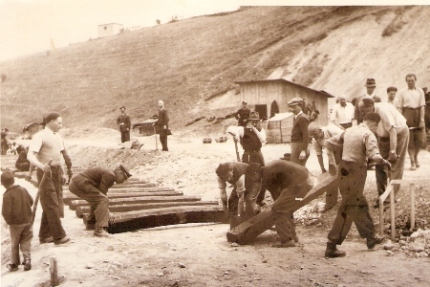  Skupina robotníkov pri kladení drevených podvalov na pláň (zemina). V pozadí drevený prístrešok. Text: Trať Turňa - Rožňava, výstavba trate. Anon. cca 1950. 170 x 123 