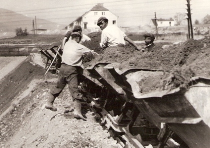  Štyria robotníci pri vyklápaní plech. huntíkov so zeminou. V pozadí normálnerozchodná trať a obytné domy. Text: Trať Turňa - Rožňava, výstavba trate. Anon., cca 1955. 172 x 12 