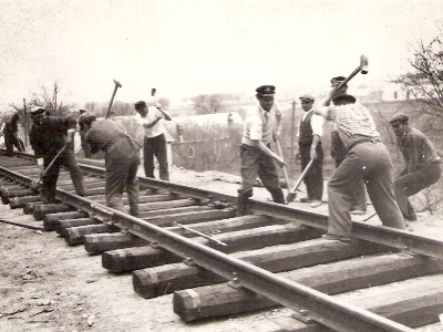  Skupina robotníkov (niektorí so železn. čiapkou) pri zatĺkaní klincov do podvalov. Montážna rozchodka, v pozadí obyt. dom. Text: Práce na Trati Družby. Anon., cca 1950. 110 x 85 