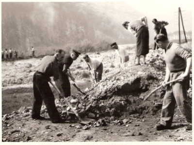  Skupina robotníkov (niektorí so železn. čiapkou) pri zemných prácach ruč. náradím v horskej krajine. V pozadí obyt. dom. Text: Trať Družby, cca 1950. 170 x 125 